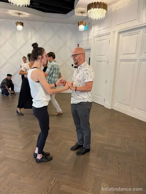 Salsa Paul demonstrating the lead with a student against the white panelled wall at The Grand Hall, Chilliwack
